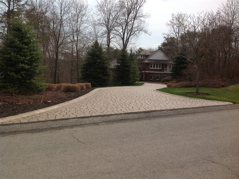Driveway of a two-story brick house with trees in the background, pavers, and a green lawn.