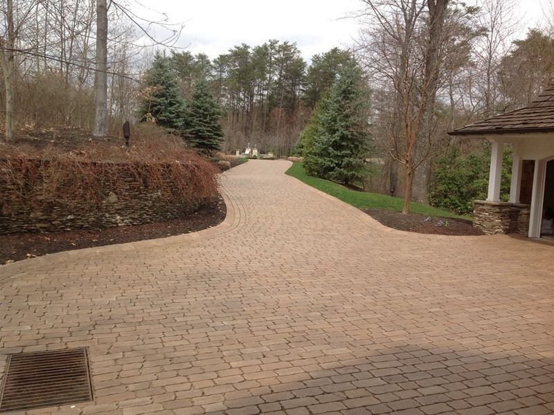 Paved brick driveway curves through landscaped yard towards trees. Brown, green, and neutral tones.