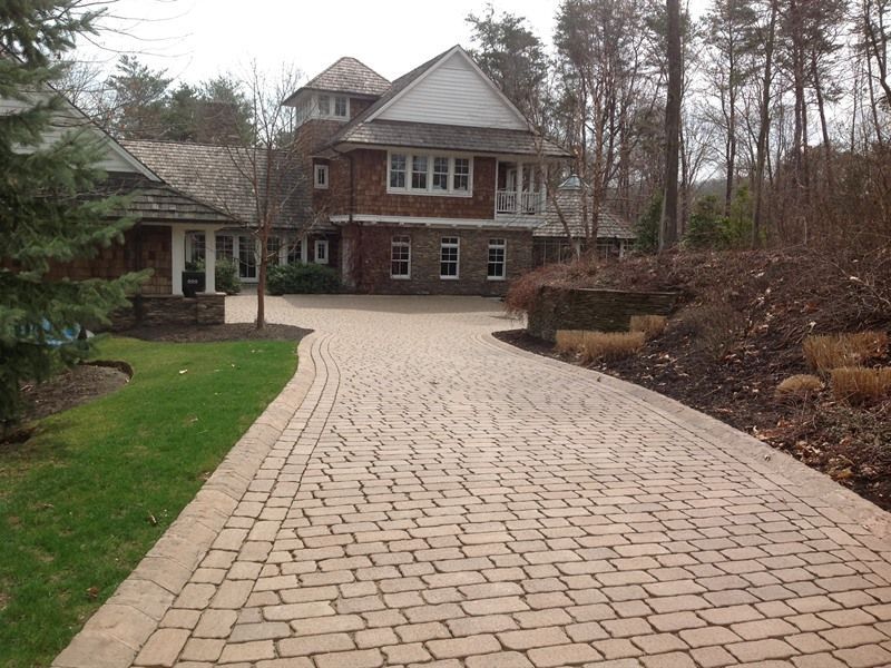 Brick driveway leading to a multi-story house with a wooden shingle roof, surrounded by trees and landscaping.