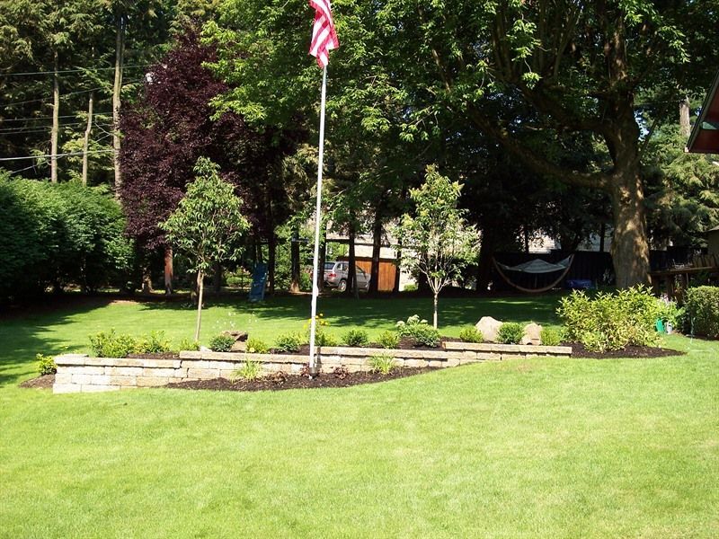 Lush green lawn with a flag pole and a stone flower bed in front of trees.