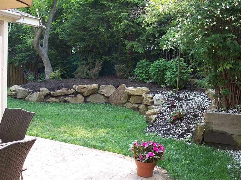 Backyard garden with stone retaining wall, green grass, and potted flowers.