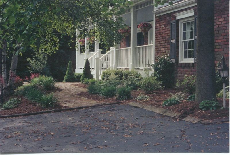 Exterior of a brick house with a white porch. Landscaped front yard with plants, mulch, and a driveway.