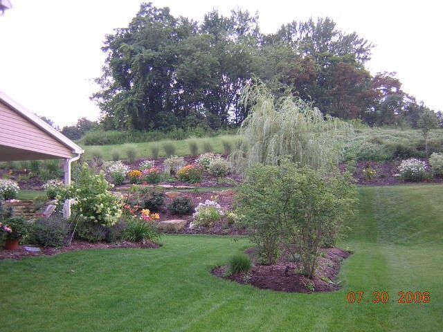 Green lawn, tiered flower beds with colorful blooms, weeping willow, trees in the background, daytime.