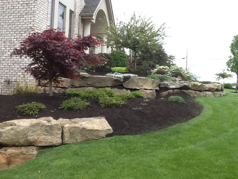Stone retaining wall with plants, mulch, and red Japanese maple tree in a grassy yard.