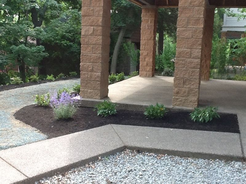 Covered outdoor area with stone columns, landscaped with plants and gravel driveway.