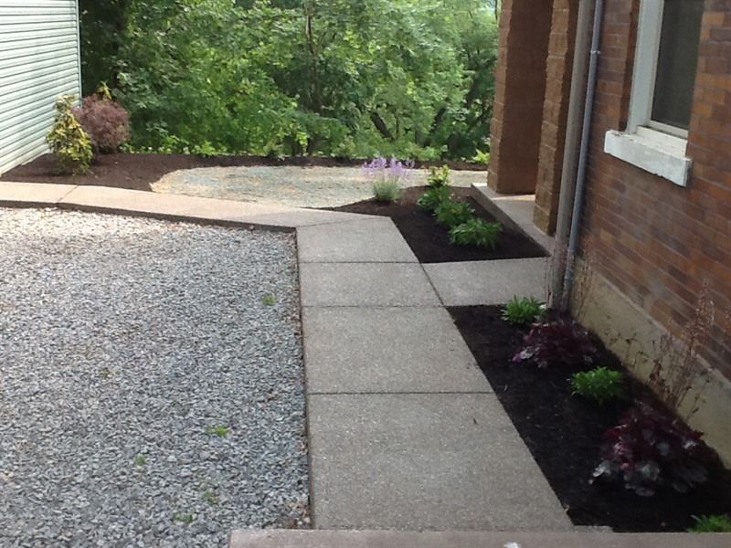 Concrete walkway leading from gravel to a building entrance with flower beds and trees.