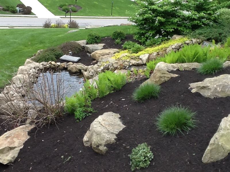 A landscaped rock garden with a small pond, surrounded by lush green plants and dark mulch.