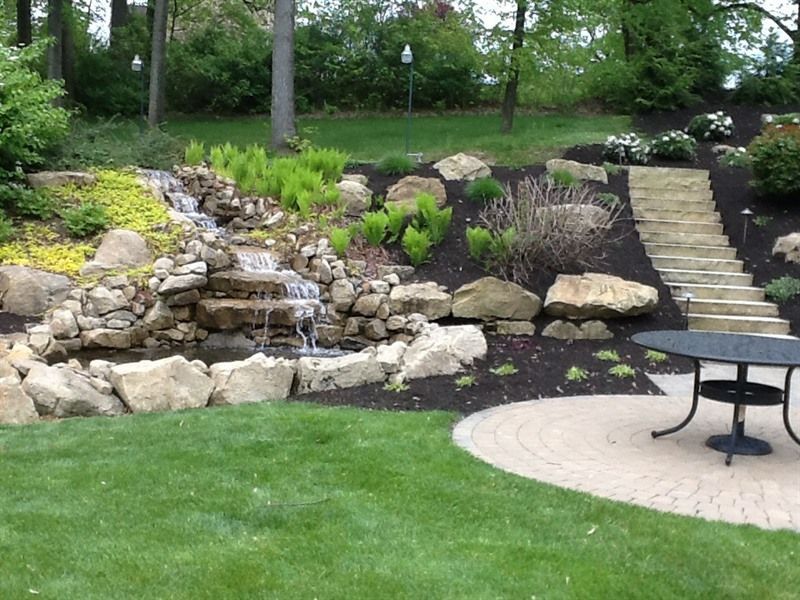 Waterfall feature with tiered landscaping, stone steps, and a patio area with table.