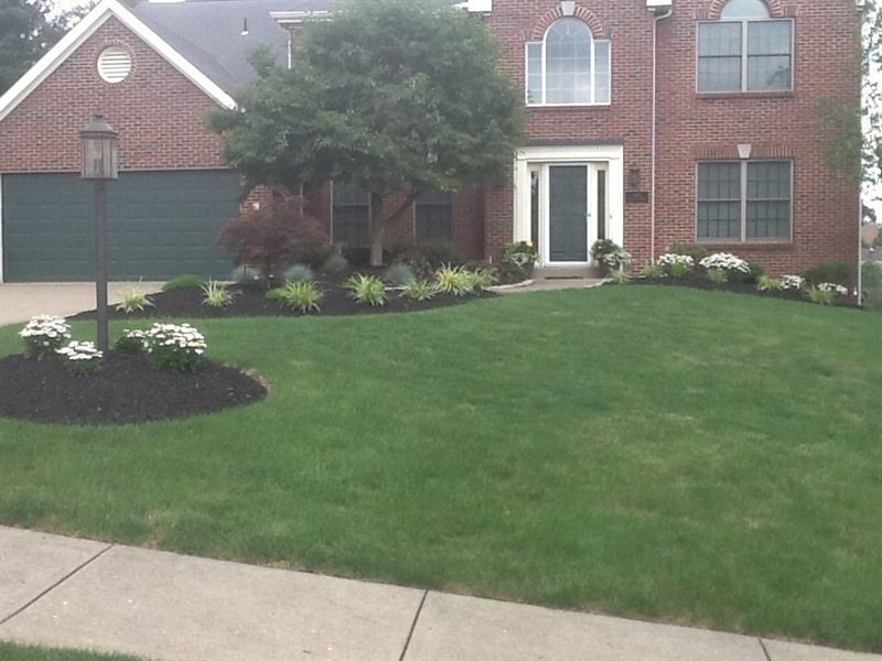Two-story brick house with green lawn, flower beds, and dark mulch. A black lamp post is visible.