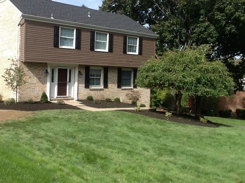 Two-story house with brown siding, black shutters, brick facade, and a well-manicured lawn and landscaping.
