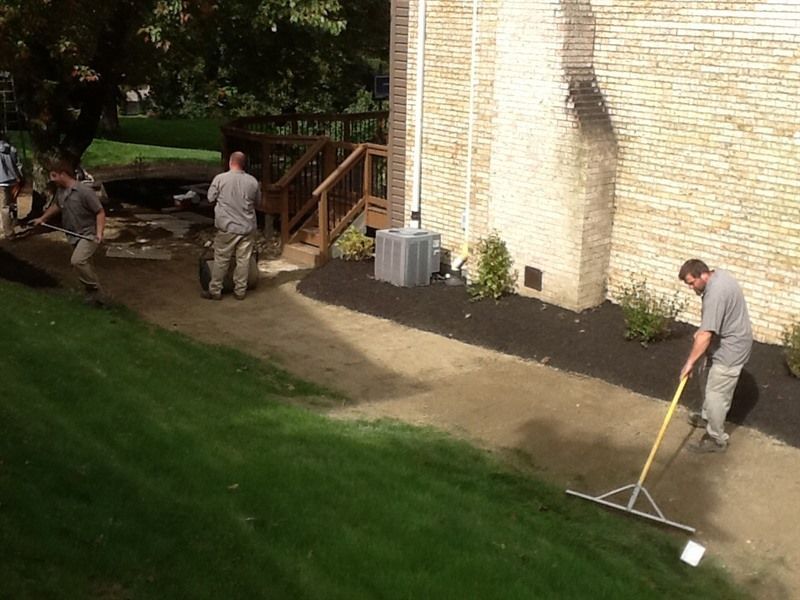Workers landscaping a walkway next to a building. One rakes gravel; others work on a deck and pathway.
