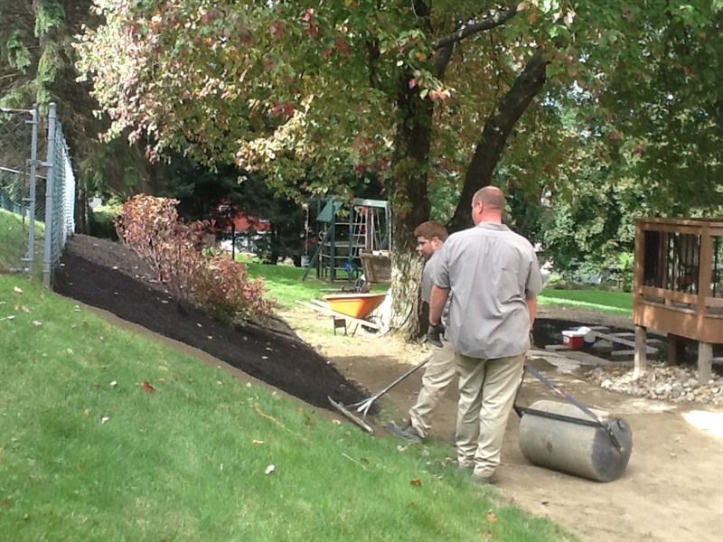 Two people raking mulch on a slope near a playground and wooden structure, with a rolled mat visible.