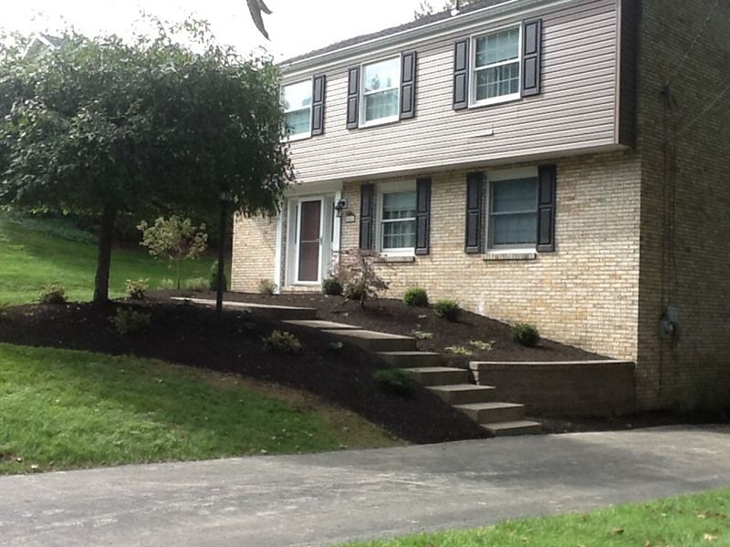 Two-story brick house with steps leading up to the front door, landscaped with dark mulch and shrubs.