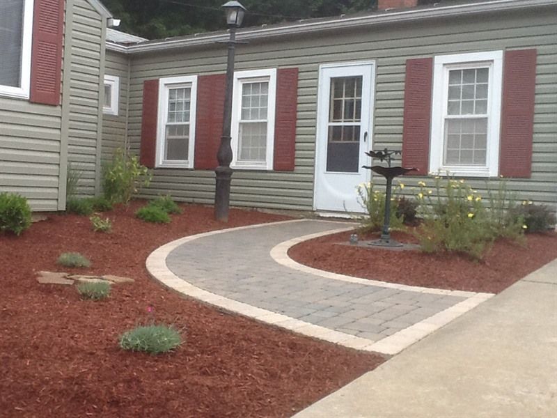 Curved brick walkway leading to a white door between two windows, framed by red shutters on a green house.