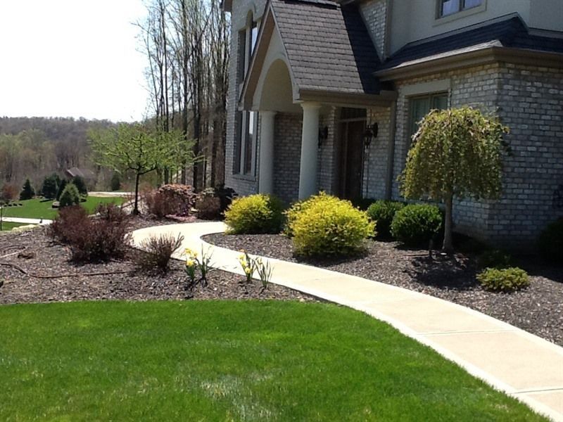 A concrete walkway curves toward a two-story house with stone and columns, flanked by landscaping and trees.
