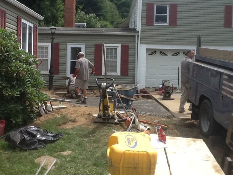 Construction workers compacting soil in a yard. Buildings and a truck are visible.