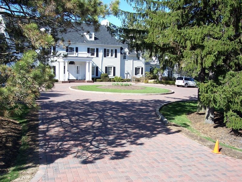 White mansion with circular driveway and brick path, trees, and blue sky.