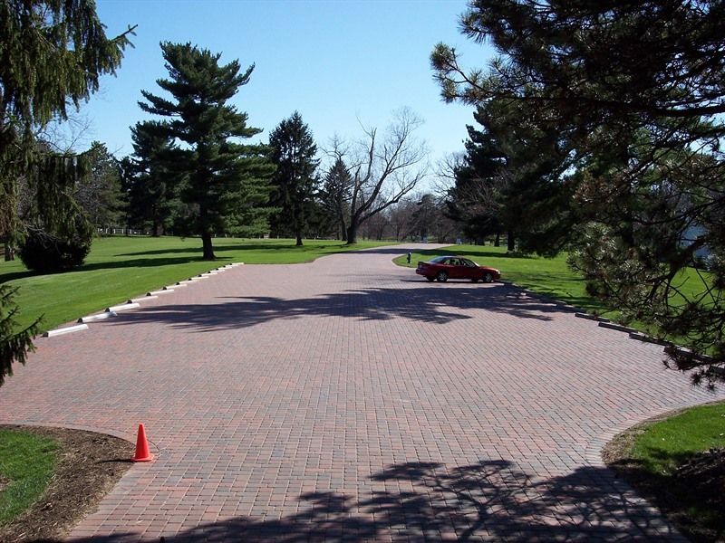 Brick driveway curves into the distance; red car parked on the right. Green grass, trees, and blue sky.