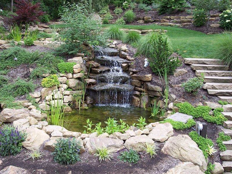 Small waterfall cascading into a pond, surrounded by rocks and lush greenery on a terraced hillside.