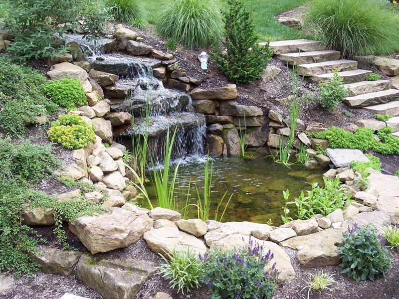 A waterfall cascading into a pond, surrounded by rocks, steps, and lush greenery.