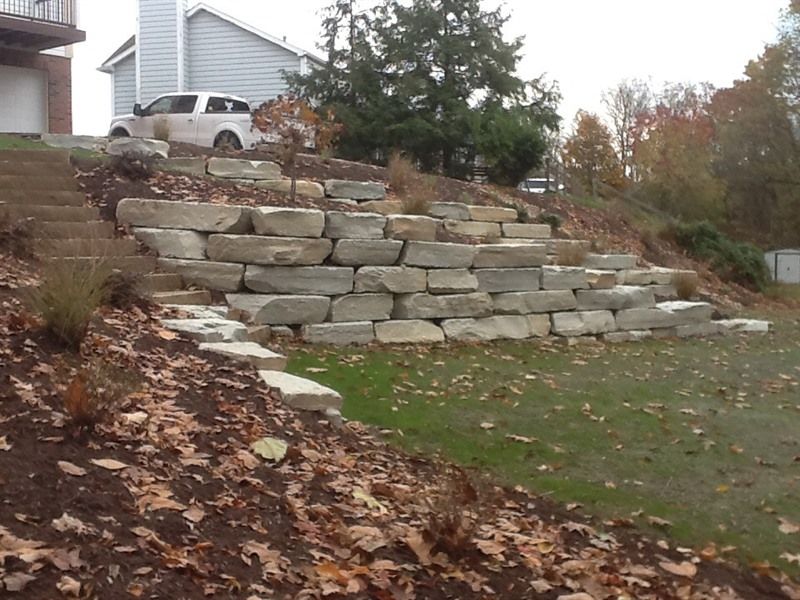 Stone retaining wall with steps, on a sloped yard next to a house. Autumn foliage and a truck are visible.