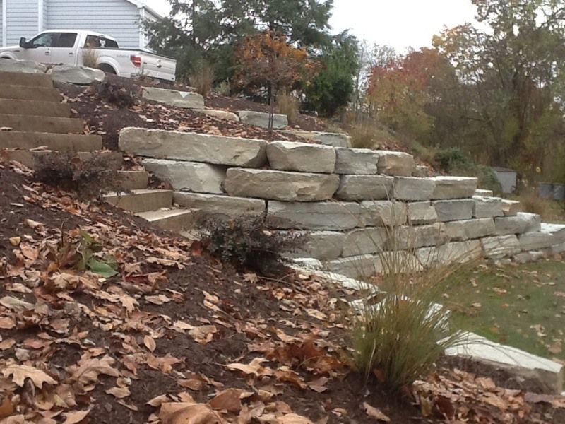 Stone retaining wall on a hillside with steps, surrounded by plants and autumn leaves.