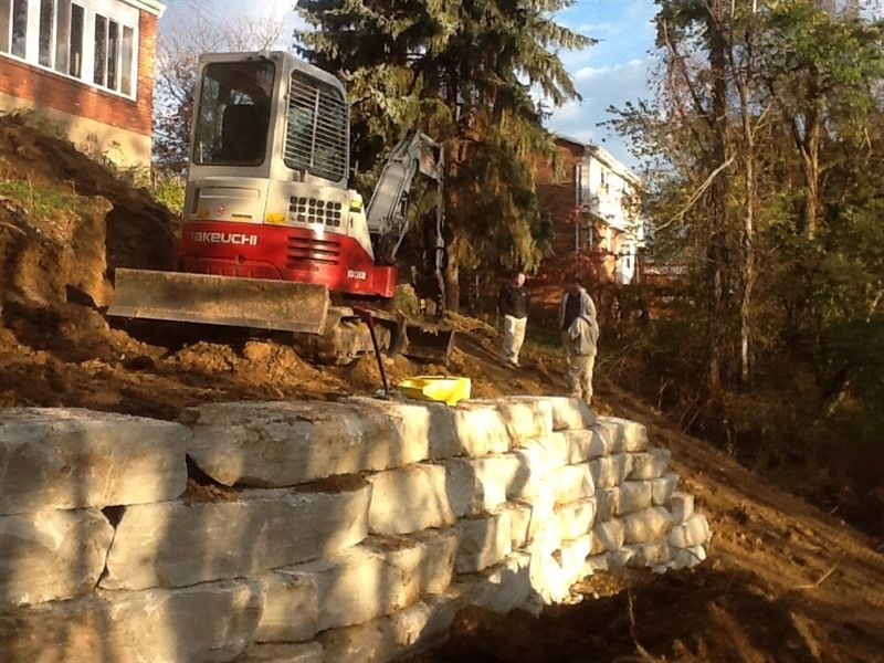 Mini excavator on a hillside constructing a retaining wall of stacked concrete blocks; a person stands nearby.