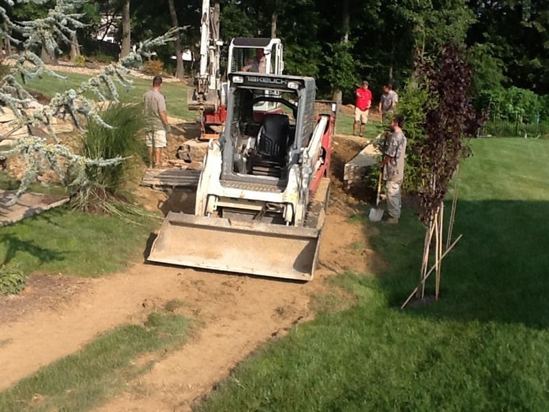 Construction crew using a Bobcat and drill on a dirt path next to green grass.
