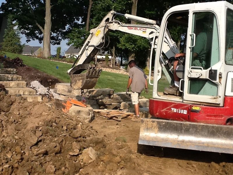 Mini excavator removing rocks from a hillside with a man working nearby. Sunny outdoor setting.