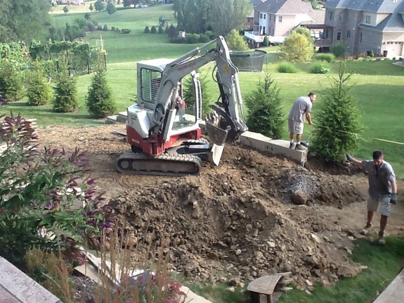 Excavator digging in backyard, two workers nearby; residential area, sunny day.