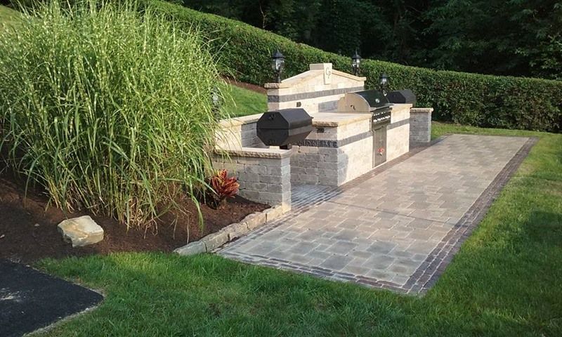 Outdoor kitchen with stone grill and path, surrounded by grass and tall ornamental grass.