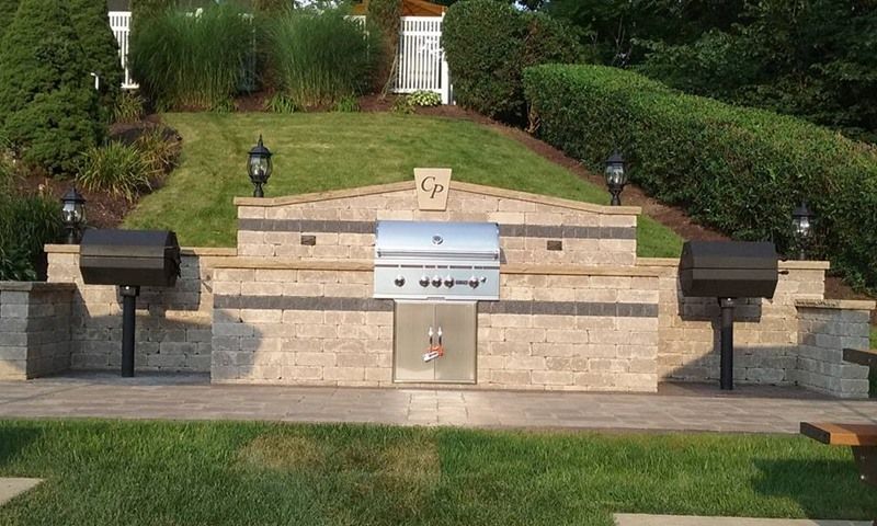 Outdoor kitchen with built-in grill, side burners, and stone structure on a green lawn.