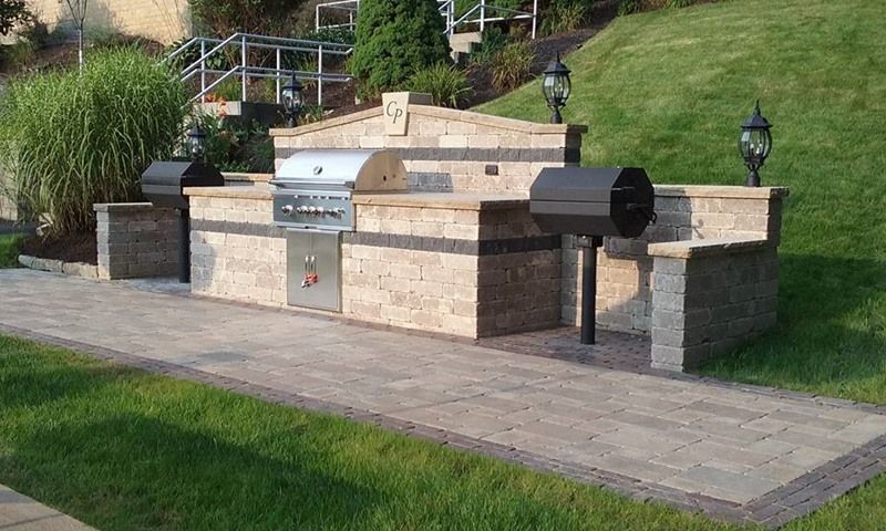 Outdoor kitchen with built-in grill, smoker, and countertops made of stone, set on a stone patio with lawn.