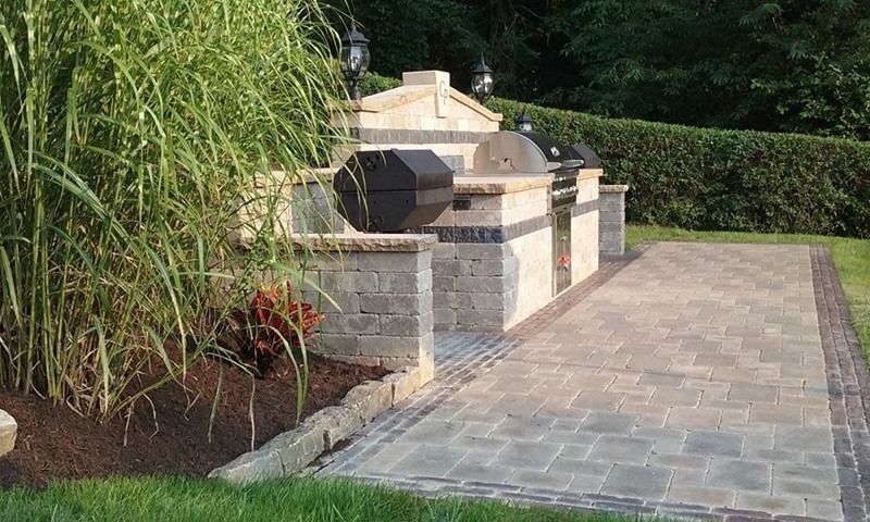 Stone outdoor kitchen with grill on paved patio, with tall grass and foliage nearby.