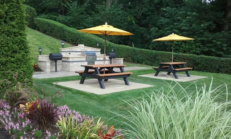 Picnic area with two tables, yellow umbrellas, and a grill. Lush green surroundings.