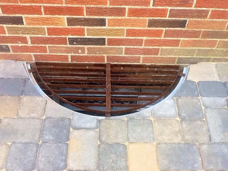Brick wall above a half-circle window well with a metal grate, set into a paved walkway.