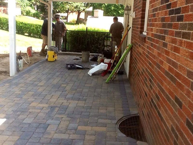 Brick patio with workers; tools and materials scattered. Side of brick building, patio under a white pergola.
