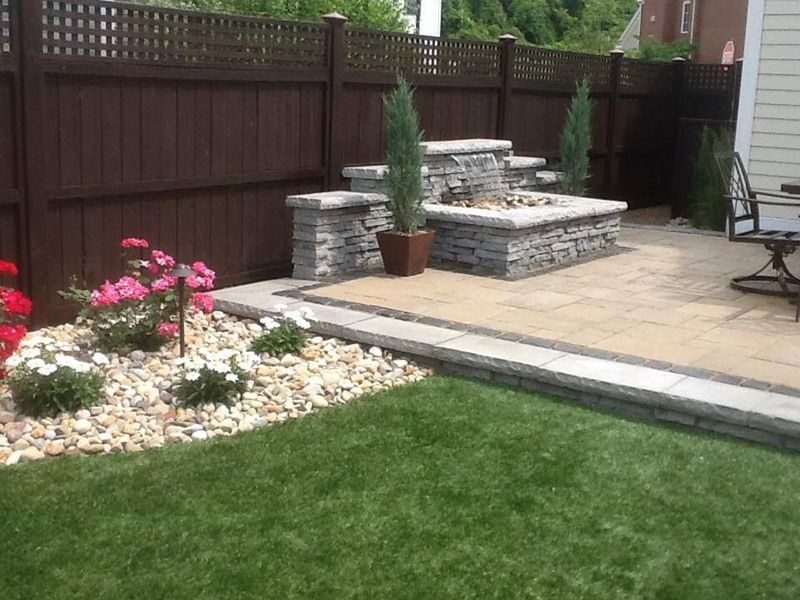 Backyard patio with stone water feature, brown fence, flowerbed, and green grass.