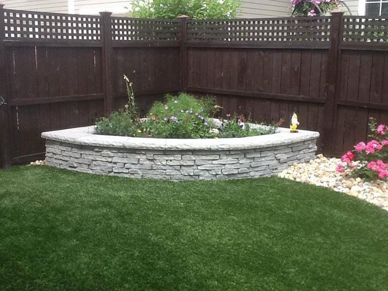 Stone planter bed in a corner of a brown wooden fenced yard, with green grass and flowers.