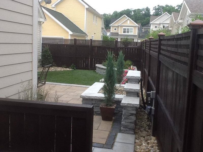Brown fence surrounds a small backyard with a patio, stone walls, and small evergreens.
