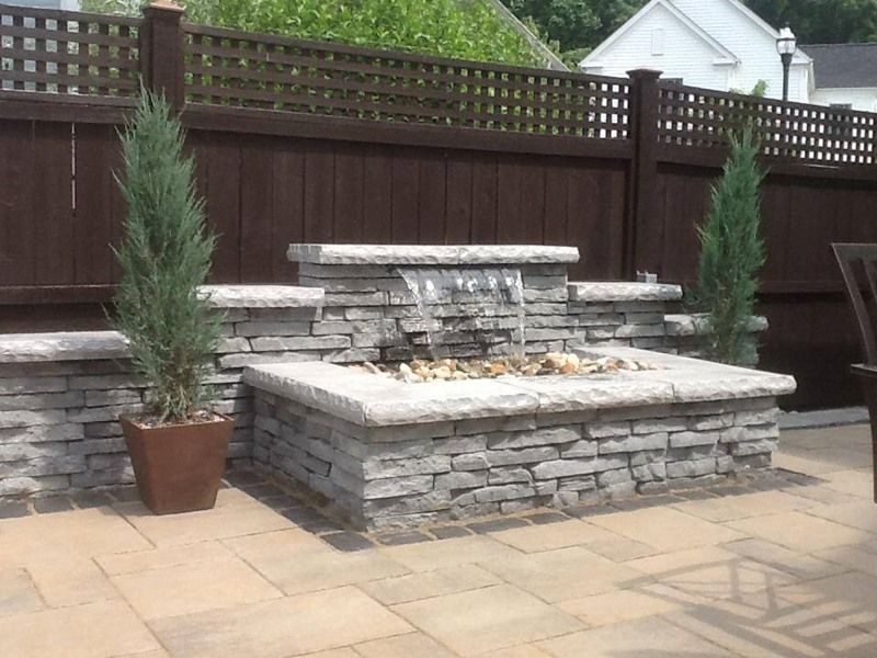 Stone water feature in a backyard, with waterfall, two potted trees, and brown fence.