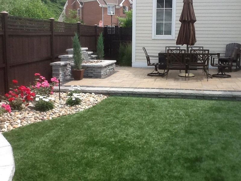 Lush green lawn with patio and fire pit area. Brown fence, stone steps, and dining set.