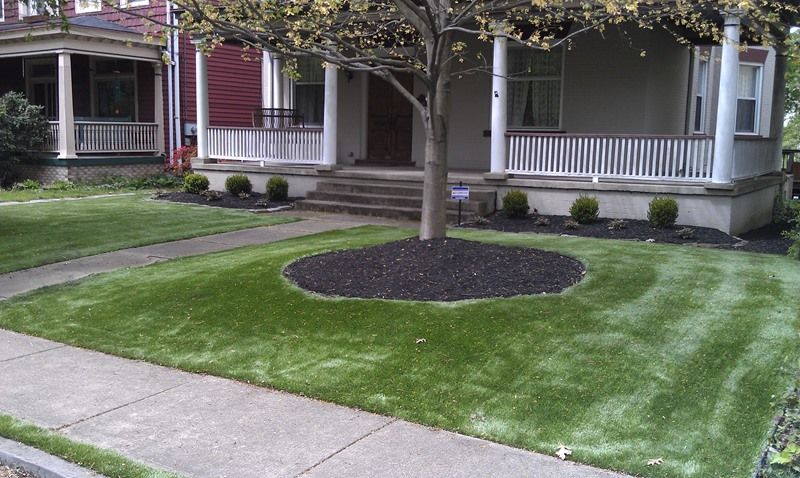 A house with a porch, a tree in the yard surrounded by mulch, and a sidewalk.