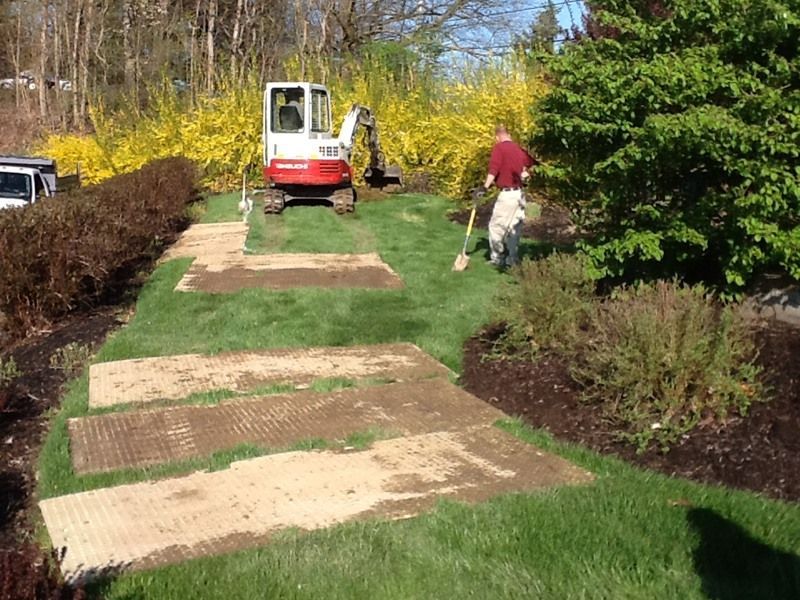 A person and excavator prepare a garden bed. Patches of grass removed, surrounded by bushes.