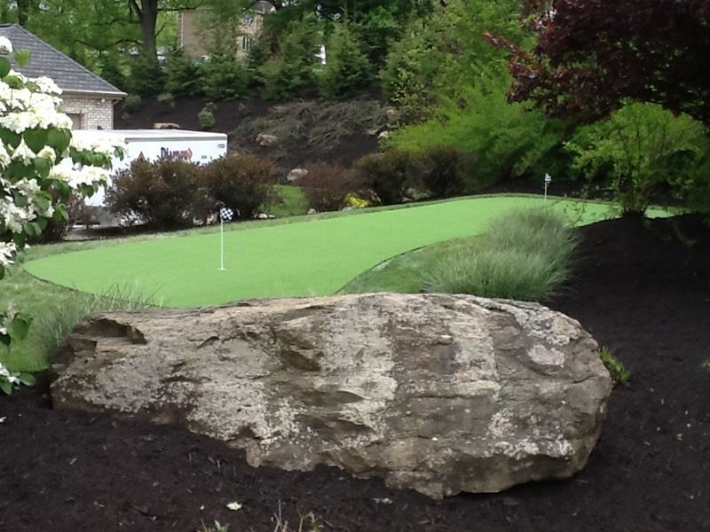 A backyard putting green with a large rock in the foreground, surrounded by landscaping.