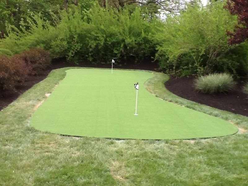 A backyard putting green with two flags, surrounded by landscaping and lawn.