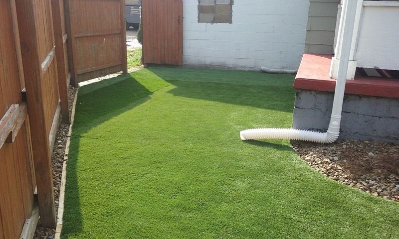 A small backyard with green artificial turf, brown fence, white building, and downspout.