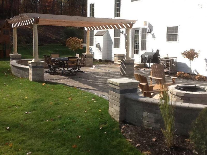 Patio with pergola, seating area, and fire pit in a backyard with lawn and white house.