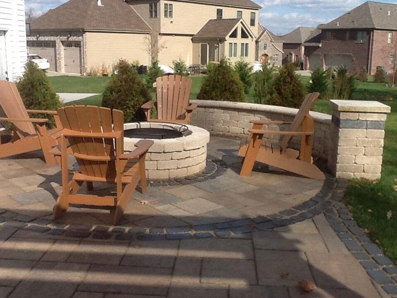 Patio with fire pit surrounded by chairs. Brick homes and green lawns in background.