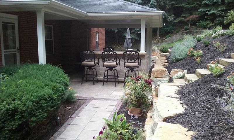 Outdoor bar with barstools on patio near a brick house. Steps and plants on a hillside.